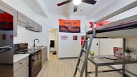 This compact studio is a single-floor unit thoughtfully designed to maximize functionality within a modest footprint. The space is organized into three distinct zones: a living/sleeping area, a kitchenette, and a private bathroom.

The main living area features light hardwood-style flooring, white walls, and a ceiling fan that contributes to a bright, airy feel. A loft-style bed frame with an integrated ladder and study desk beneath makes excellent use of vertical space, offering both a sleeping area and a dedicated workspace without compromising floor area.

The kitchenette is efficiently outfitted with gray shaker-style cabinetry — both upper and lower — complemented by light countertops. Appliances include a black electric range and a microwave, along with a single-basin sink with a modern matte black faucet. This kitchen area provides all the essential amenities for everyday meal preparation.

The private bathroom, accessed through a separate door, features a toilet and compact layout with tiled flooring. Exposed painted cinder block walls in a secondary area of the unit add an industrial character to the overall aesthetic.

A window allows natural light into the space, and the unit includes an entry door painted in a bold black finish. Overall, the studio presents a practical and functional layout well-suited for a single occupant seeking a self-contained living environment.