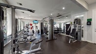 This fitness center is a fully equipped, single-floor exercise facility designed to support a wide range of workout routines. The space is thoughtfully laid out, with dedicated zones for both cardiovascular training and strength work. Cardio equipment includes multiple elliptical machines and treadmills, strategically positioned along a wall of large windows that frame views of lush greenery, creating an energizing and open atmosphere during workouts. Mounted flat-screen TVs throughout the space provide entertainment while exercising.

The strength training area is well-appointed with cable and pulley machines, a multi-functional cable crossover unit, adjustable benches, and a dedicated free weight section featuring a full dumbbell rack. Additional equipment such as medicine balls and body weight benches further expand workout versatility.

Floor-to-ceiling mirrors line multiple walls, enhancing the sense of space and allowing users to monitor their form. Recessed lighting illuminates the facility evenly, complemented by generous natural light from the expansive windows. The flooring consists of durable dark rubber matting, providing comfort and safety underfoot. The overall aesthetic is clean and modern, with a neutral white and grey palette that contributes to a focused, professional training environment.