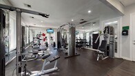 This fitness center is a fully equipped, single-floor exercise facility designed to support a wide range of workout routines. The space is thoughtfully laid out, with dedicated zones for both cardiovascular training and strength work. Cardio equipment includes multiple elliptical machines and treadmills, strategically positioned along a wall of large windows that frame views of lush greenery, creating an energizing and open atmosphere during workouts. Mounted flat-screen TVs throughout the space provide entertainment while exercising.

The strength training area is well-appointed with cable and pulley machines, a multi-functional cable crossover unit, adjustable benches, and a dedicated free weight section featuring a full dumbbell rack. Additional equipment such as medicine balls and body weight benches further expand workout versatility.

Floor-to-ceiling mirrors line multiple walls, enhancing the sense of space and allowing users to monitor their form. Recessed lighting illuminates the facility evenly, complemented by generous natural light from the expansive windows. The flooring consists of durable dark rubber matting, providing comfort and safety underfoot. The overall aesthetic is clean and modern, with a neutral white and grey palette that contributes to a focused, professional training environment.