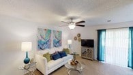 This single-floor residence offers a comfortable and stylishly appointed living space designed for everyday ease and relaxed living. The living room serves as the heart of the home, featuring plush wall-to-wall carpeting that adds warmth underfoot and a ceiling fan with integrated lighting to maintain a comfortable atmosphere year-round. A large white sofa anchors the seating area, complemented by a glass-topped coffee table and coordinating accent furniture that lend the space a modern, airy feel. Vibrant abstract artwork in teal, purple, and gold tones adorns the walls, infusing the room with color and personality. A media console with decorative lattice detailing provides practical storage alongside the flat-screen television. Teal drape panels frame a window that draws in natural light, brightening the neutral-toned walls and textured ceiling. Decorative wall accents and carefully chosen accessories throughout contribute to a cohesive, curated aesthetic. The overall ambiance is inviting and contemporary, blending functional layout with thoughtful design details that make the space feel both livable and visually engaging.