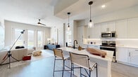 This thoughtfully designed 2-bedroom, 3-bathroom residence spans a single floor and offers a modern, open-concept layout with light wood-tone flooring and a neutral color palette throughout. The combined kitchen and living area forms the heart of the home, anchored by a sleek white kitchen featuring shaker-style cabinetry extending to the ceiling, stainless steel appliances, under-cabinet lighting, and a large center island with a waterfall countertop and bar seating for two, illuminated by a pair of glass pendant lights. The living room is bright and airy, benefiting from abundant natural light through full-height glass doors that open to a private balcony with open green views. The primary bedroom is a spacious retreat with elevated ceilings, direct access to a dedicated en-suite bathroom with a dual-sink vanity, black-framed mirrors, and dark hardware accents, plus a walk-in closet with a custom shelving and drawer organization system. The second bedroom is well-proportioned and includes a nearby bathroom with a double vanity. A dedicated laundry room with a full-size washer and ample shelving, two closets, and a connecting hallway round out this well-organized and functional floor plan.