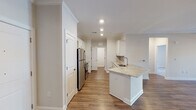Located at 5300 High Point Rd in Greensboro, NC, this single-floor unit offers a well-designed layout with a clean, modern aesthetic throughout. The kitchen is a standout feature, showcasing white shaker-style cabinetry with brushed nickel hardware, light granite countertops, a subway tile backsplash, and stainless steel appliances including a refrigerator and range. A peninsula with a built-in sink provides additional workspace and casual dining potential. Wood-look flooring runs through the main living and kitchen areas, adding warmth and continuity. The open living area is bathed in natural light and features crown molding for an elevated finish. Bedrooms are carpeted for comfort, each equipped with a ceiling fan and window with blinds; one bedroom offers French doors that open to an outdoor view. The primary bathroom includes a long dual-sink vanity with granite countertops, a separate walk-in shower, and wood-look flooring. A secondary bathroom provides a full tub and shower combo with clean white surround. A dedicated laundry room houses a full-size side-by-side washer and dryer with overhead shelving, adding everyday convenience. The unit presents a fresh, move-in-ready feel throughout.