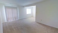 The unit at 27 Shawnee Ct 302 is a single-floor residence characterized by its open and airy layout. The interior features wall-to-wall beige carpeting throughout, providing a warm, cohesive feel across the living spaces. The main living area is generously sized, with white walls and a standard ceiling height that contribute to a bright, clean atmosphere. Vertical blinds on the left side of the living area cover what appears to be a sliding door or large window, offering both privacy and light control. An open passageway connects the main living space to an adjacent room, which benefits from a centered window that allows natural light to filter in, further enhancing the overall brightness of the unit. A ceiling-mounted light fixture in the secondary room provides additional illumination. The neutral color palette throughout — from the off-white walls to the light carpeting — creates a versatile backdrop that easily accommodates a variety of furnishing styles. The space presents a clean, move-in ready condition with a straightforward, functional layout suited for everyday living.