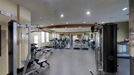 This fitness center offers a thoughtfully designed single-floor layout that accommodates a diverse selection of exercise equipment. The space features a generous open floor plan with a smooth, durable dark flooring surface suited for high-traffic use. Recessed lighting throughout the ceiling provides bright, even illumination across the entire area, creating a functional and energizing atmosphere.

Along one side of the room, a dedicated free weights section includes a full rack of dumbbells alongside adjustable weight benches, catering to strength training needs. The central portion of the floor is lined with multiple cardio machines, including treadmills, ellipticals, and upright stationary bikes, offering ample options for aerobic exercise. A cable and pulley machine system is positioned along the perimeter, providing additional resistance training versatility.

The ceiling features an architectural curved accent beam that adds a subtle design element to the otherwise clean, utilitarian aesthetic. Windows along one wall allow natural light to filter in, complementing the recessed overhead lighting. The overall layout is open and well-organized, allowing for comfortable movement between equipment stations.