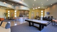 This contemporary community lounge is designed to offer a dynamic and inviting environment for recreation and socializing. The open-plan layout seamlessly connects distinct activity zones, creating a fluid and spacious feel throughout. A standout feature is the elegant curved architectural detail on the ceiling, which adds a sculptural element to the room and contributes to its modern character.

At the center of the space sits a sleek, dark-framed pool table with a white felt surface, positioned atop a textured area rug that defines the recreation zone. Overhead, a stylish multi-pendant light fixture with copper-toned accents provides focused illumination over the table.

The kitchen area is well-appointed, featuring dark cabinetry, a mosaic tile backsplash, and a stainless steel range hood as a focal point. A long bar-height counter with vibrant orange upholstered stools runs alongside, creating a casual dining and gathering spot. Recessed lighting throughout the ceiling ensures the space is well-lit, while windows along the walls allow natural light to filter in.

Warm yellow-toned walls and polished flooring complete the energetic yet welcoming ambiance of this multifunctional lounge.
