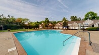 Located at 1631 Hill Rd N in Pickerington, Ohio, this single-story property is notable for its well-appointed outdoor amenities set within a verdant, tree-lined environment. The standout feature visible from the imagery is a generously sized outdoor swimming pool with clear, blue water and a smooth concrete surround. The pool deck is equipped with lounge chairs and shaded seating areas, including umbrella-covered tables, making it well-suited for outdoor relaxation and recreation. A metal safety fence encloses the pool area, providing both security and definition to the space. The surrounding landscape features mature trees and maintained greenery, contributing to a pleasant and private outdoor atmosphere. The adjacent residential structures display a traditional architectural style with neutral siding and pitched rooflines, complementing the overall character of the community. The open-sky setting allows for ample natural light throughout the outdoor area, enhancing its appeal during warmer months. The combination of the expansive pool, dedicated lounge areas, and lush surroundings creates a cohesive and functional outdoor environment well-suited for leisure and community use.