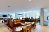 Located at The Ward in Everett, MA, this expansive amenity floor offers a thoughtfully designed, multi-zone communal space that blends modern aesthetics with warm, inviting interiors. Wide-plank light oak hardwood flooring flows throughout the open layout, unifying distinct areas while allowing each zone to maintain its own character.

The main lounge is anchored by rich cognac leather sectional sofas, layered over bold patterned area rugs, and accented with warm table lamps and curated artwork. A long bar counter with seating lines the perimeter, bathed in natural light from oversized floor-to-ceiling windows that frame views of the surrounding Everett streetscape. An adjacent dining and bar area features rows of dark wood bar stools set against a full-length window wall.

A dedicated game room showcases a central billiards table beneath a brass pendant light, complemented by a wall-mounted TV, vibrant blue sofa, and large sliding glass doors that invite natural light. A more intimate lounge area features a coffered ceiling, sage-toned paneled walls, a tufted charcoal Chesterfield sofa, and generous windows overlooking the neighborhood.

Additional spaces include a cozy library lounge with built-in dark shelving, a sputnik chandelier, and green marble coffee tables, as well as a bright, airy sitting room with black-framed windows and sculptural furnishings. A central hub area with a marble-topped island serves as a gathering and service point, tying all zones together seamlessly.