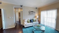 This single-story residence at WestRidge Apartment Homes offers a well-appointed living space designed with both comfort and functionality in mind. The living room serves as the heart of the home, showcasing rich dark hardwood-style flooring that contrasts elegantly with the neutral-toned walls accented by classic crown molding. A large sliding glass door dressed with vertical blinds allows natural light to fill the room, creating a bright and welcoming atmosphere. The space comfortably accommodates a full-sized sofa and a glass-top coffee table, illustrated here over a teal geometric area rug that adds a pop of color to the room. Overhead, a ceiling-mounted HVAC vent ensures efficient climate control throughout. An open doorway leads to a hallway that connects to additional rooms, with a glimpse of a well-organized closet visible beyond. The overall ambiance is clean, modern, and inviting, reflecting a thoughtfully designed floor plan that balances open living areas with practical storage and private spaces throughout the single-level layout.