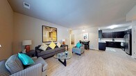 This single-floor residence features an open-concept layout that connects the living, dining, and kitchen areas into one cohesive, flowing space. The living area is anchored by a dark upholstered sofa adorned with colorful accent pillows in yellow and teal, complemented by two striped armchairs and a sleek glass-top coffee table. Light wood-style flooring extends throughout, enhancing the sense of openness and contributing to a bright, airy ambiance. Neutral gray walls provide a clean backdrop, while a large abstract wall art piece in yellow and gray tones adds a bold visual focal point. The dining area, situated just beyond the living space, features a compact table with dark chairs and is lit by a pendant light overhead. Adjacent to the dining area, the kitchen is partially visible and outfitted with dark cabinetry and light countertops. A hallway leading to additional rooms is visible in the background, suggesting a well-organized floor plan. Overall, the space presents a modern, well-appointed interior with a cohesive color palette and functional open design.