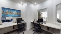 This single-floor business center offers a practical and efficiently designed workspace tailored for productivity. The room features two dedicated workstations arranged along built-in white desks that wrap around adjacent walls, each equipped with a desktop computer, keyboard, and ergonomic mesh office chair. The layout maximizes the available space while ensuring each station feels distinct and functional.

The interior is finished with neutral white walls and carpeted flooring, contributing to a clean, professional atmosphere. Recessed ceiling lights provide consistent, even illumination throughout the space, complemented by a textured ceiling that adds subtle visual interest. A colorful cityscape painting on one wall introduces a vibrant accent, enhancing the otherwise minimalist aesthetic.

A large pass-through window or open partition connects this workspace to an adjacent, naturally lit room, which appears to feature dining furniture and benefits from outside light — lending an open, airy quality to the overall environment. A wall clock is visible through the opening, adding a practical touch. The space is well-suited for shared or co-working use in a residential or boutique professional setting.