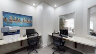 This single-floor business center offers a practical and efficiently designed workspace tailored for productivity. The room features two dedicated workstations arranged along built-in white desks that wrap around adjacent walls, each equipped with a desktop computer, keyboard, and ergonomic mesh office chair. The layout maximizes the available space while ensuring each station feels distinct and functional.

The interior is finished with neutral white walls and carpeted flooring, contributing to a clean, professional atmosphere. Recessed ceiling lights provide consistent, even illumination throughout the space, complemented by a textured ceiling that adds subtle visual interest. A colorful cityscape painting on one wall introduces a vibrant accent, enhancing the otherwise minimalist aesthetic.

A large pass-through window or open partition connects this workspace to an adjacent, naturally lit room, which appears to feature dining furniture and benefits from outside light — lending an open, airy quality to the overall environment. A wall clock is visible through the opening, adding a practical touch. The space is well-suited for shared or co-working use in a residential or boutique professional setting.