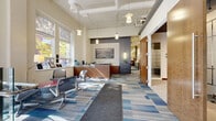 This single-floor professional office space is designed to balance functionality with a polished, contemporary aesthetic. The reception area greets visitors with soaring ceilings, pendant lighting, and a prominent arched entryway that floods the space with natural light. A warm wood reception desk anchors the area, complemented by modern seating, a decorative area rug, and a framed aerial cityscape on the slate-blue accent wall.

The open workspace extends deeper into the office, featuring rows of modular workstations with partial-height dividers, providing individual focus areas while maintaining an open, collaborative atmosphere. Exposed ductwork and structural elements add an industrial character to the high ceilings, while linear fluorescent lighting ensures consistent illumination throughout.

Multiple enclosed conference rooms offer versatile meeting options. One light-filled room features expansive windows with outdoor tree views and a large wood conference table, while a second, more formal room is equipped with a wall-mounted display screen and track lighting set against a dark ceiling. Private offices with glass-panel sliding doors and wood accents provide additional enclosed workspace.

Throughout the office, a consistent blue, grey, and cream patterned carpet tile unifies the various zones, contributing to a cohesive and professional overall ambiance.