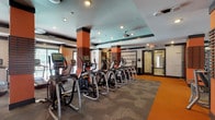 Located at 3853 Forest Park Ave in St. Louis, Missouri, this fitness center is a thoughtfully designed, single-floor facility offering a comprehensive workout environment. The space features a striking interior palette of bold orange, dark charcoal, and neutral tones, with distinctive orange structural columns wrapped in horizontal wood slat detailing that serve as architectural focal points throughout.

The cardio zone is well-appointed with a row of elliptical trainers, treadmills, and other aerobic machines, each equipped with individual digital displays. Patterned geometric carpeting covers the main workout floor, while vibrant orange rubber flooring delineates key circulation paths and zones, adding both visual energy and functional wayfinding.

The strength training area houses a full selection of cable machines, weight benches, free weights, and multi-station equipment, all arranged to allow comfortable movement between stations. Large mirrors are strategically placed to maximize the sense of space and support proper exercise form.

A dedicated studio or flex space features wall-mounted flat-screen televisions, a digital kiosk, exercise balls, and step platforms, suitable for group classes or individual training sessions. Natural light filters in through large windows, offering views of the surrounding outdoor environment. Recessed and ceiling-mounted lighting ensures the entire facility is well-illuminated throughout.