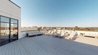 Located atop 165 Huguenot Street in New Rochelle, New York, this rooftop terrace is a well-appointed outdoor amenity space offering sweeping, unobstructed views of the New Rochelle skyline, neighboring architecture, and tree-lined surroundings. The terrace is finished with wide-plank gray composite decking that extends generously across the open layout, providing a clean, modern aesthetic underfoot.

The space is furnished with multiple cushioned chaise lounges arranged to take full advantage of the sun exposure, along with a substantial sectional sofa seating area ideal for relaxed gatherings. The combination of lounging and seating zones allows the terrace to accommodate both quiet relaxation and social interaction.

Access to the terrace is provided through a set of floor-to-ceiling bronze-framed glass doors set into a crisp white paneled facade, allowing a seamless visual connection between interior and exterior spaces. Low parapet walls line the perimeter, maintaining open sightlines to the surrounding cityscape, which includes prominent high-rise towers and the wider New Rochelle urban fabric. The terrace benefits from abundant natural light and open sky, making it a highly functional and visually striking outdoor amenity.