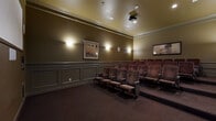 This private screening room offers a complete theater experience within a compact, thoughtfully designed space. The room is arranged with multiple rows of plush, cushioned theater-style seats in a warm mauve upholstery, each equipped with built-in armrests and cup holders for added comfort. The seating is tiered to ensure unobstructed sightlines toward a large projection screen mounted on the front wall, with a ceiling-mounted projector positioned overhead for optimal image display. The walls are finished in a rich olive-green tone, accented by classic wainscoting panels that lend a refined, traditional aesthetic. Crown molding runs along the ceiling perimeter, enhancing the room's polished character. Soft wall sconces provide warm, ambient lighting, complemented by framed landscape artwork displayed throughout the room. Deep, dark carpeting covers the entire floor, contributing to both the acoustic quality and the intimate atmosphere of the space. A charming red popcorn machine stationed along the rear wall adds a playful, nostalgic touch reminiscent of a classic movie house. A paneled entry door provides access, completing this well-appointed, self-contained cinema room.