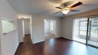 This single-story space is designed with a practical, open layout that connects the main living and dining areas seamlessly. Dark hardwood-style flooring runs throughout the common areas and hallway, providing a warm, consistent aesthetic, while plush carpet adds comfort to the bedrooms. Textured ceilings are present throughout, and ceiling fans with integrated lighting fixtures are installed in the living areas and bedrooms, contributing to both air circulation and ambient illumination.

The living area is generously proportioned, featuring vertical blinds along a sliding door or large window that allows natural light to filter in. A pass-through window connects the living and dining space to the kitchen, enhancing the open feel. The kitchen is well-appointed with white cabinetry, granite-style countertops, a stainless steel refrigerator, and a tile backsplash.

The bedrooms are carpeted and well-lit by natural light from multiple windows fitted with blinds. At least one bedroom includes a closet with built-in shelving. The bathroom features a granite-top vanity with a single sink, a tub-shower combination with tiled surround, and dark hardwood-style flooring, maintaining cohesion with the rest of the space.