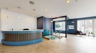 Located at 163 Franklin St in Stamford, Connecticut, this professionally designed commercial space offers a polished and welcoming environment ideal for client-facing operations. The open-plan layout is anchored by a striking curved reception desk accented with wood-stripe detailing and a bold teal base, seamlessly blending functionality with contemporary style. Underfoot, light-toned herringbone hardwood floors extend throughout, adding warmth and visual depth to the space.

The interior is defined by a rich navy and slate blue color palette across the walls, complemented by crisp white ceilings fitted with recessed lighting and decorative pendant fixtures that cast an inviting glow. A glass partition wall elegantly separates the entry and reception area from a compact kitchenette, which is outfitted with wood-grain cabinetry, a stainless steel sink, and modern appliances. Vertical blinds along large windows allow abundant natural light to pour in while maintaining privacy.

A separate seating nook, enclosed by glass and painted in deep navy, provides an intimate area for consultations or meetings, furnished with a round glass table and contemporary upholstered chairs. Tasteful wall art adds a refined finishing touch. The overall ambiance is sleek, modern, and professional, making this a highly functional space suited for a variety of business uses.