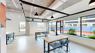 This single-floor commercial space is designed with a contemporary industrial aesthetic that balances functionality with creative energy. The open-plan layout is defined by exposed ceilings that reveal structural steel beams, ductwork, and mechanical systems, all painted in crisp white and black tones to create a visually dynamic overhead canopy. Matte concrete-style flooring runs throughout, lending the space a clean, cohesive feel underfoot.

The workspace is furnished with multiple high-top communal tables paired with modern slate-blue chairs, offering flexible configurations suited for collaboration, informal meetings, or individual work. Pendant lighting in classic black dome fixtures hangs at measured intervals, providing focused illumination that complements the ample natural light pouring in through a generous arrangement of large windows and glass doors along the perimeter. These openings frame outdoor views and bring in a bright, airy quality throughout the day.

A standout feature of the space is a large-scale, bold geometric mural adorning one full wall — an eye-catching composition of red, blue, orange, and grey shapes that serves as a vibrant focal point and injects a strong sense of artistic character. Glass-panel interior doors along another wall hint at adjacent rooms, maintaining visual connectivity. The overall ambiance is energetic yet approachable, making this space well-suited for a modern office, coworking environment, or creative studio.