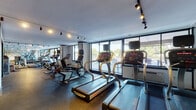 This fitness center offers a fully outfitted workout environment designed to accommodate a wide range of exercise needs. The space is organized into distinct zones, with a dedicated cardio area lining the perimeter and a spacious open floor for strength training. The cardio section features multiple treadmills, stationary bikes, and elliptical machines, all positioned along floor-to-ceiling windows that flood the area with natural light and provide open views of the outdoors. The strength training zone is generously stocked with an extensive dumbbell rack, weight benches, cable machines, and functional training equipment, supported by full-length mirrors that enhance the sense of space. Durable rubber flooring covers the entire area, providing both safety and performance underfoot. Track lighting runs throughout the ceiling, offering consistent, well-distributed illumination across all workout areas. The interior design is clean and modern, featuring geometric accent wall graphics that add a contemporary aesthetic touch. A separate room with glass partition walls houses additional strength and cable machines, offering a more focused training environment. Overall, the space maintains a professional, well-maintained atmosphere suited for a comprehensive fitness experience.