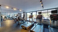 This fitness center offers a fully outfitted workout environment designed to accommodate a wide range of exercise needs. The space is organized into distinct zones, with a dedicated cardio area lining the perimeter and a spacious open floor for strength training. The cardio section features multiple treadmills, stationary bikes, and elliptical machines, all positioned along floor-to-ceiling windows that flood the area with natural light and provide open views of the outdoors. The strength training zone is generously stocked with an extensive dumbbell rack, weight benches, cable machines, and functional training equipment, supported by full-length mirrors that enhance the sense of space. Durable rubber flooring covers the entire area, providing both safety and performance underfoot. Track lighting runs throughout the ceiling, offering consistent, well-distributed illumination across all workout areas. The interior design is clean and modern, featuring geometric accent wall graphics that add a contemporary aesthetic touch. A separate room with glass partition walls houses additional strength and cable machines, offering a more focused training environment. Overall, the space maintains a professional, well-maintained atmosphere suited for a comprehensive fitness experience.