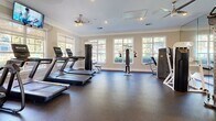Located at 1 Sycamore Ln in Woodstock, GA, this well-appointed fitness center offers a comprehensive exercise facility designed to support a full range of workout routines. The single-floor space is finished with durable rubber flooring throughout, providing a safe and functional surface for all types of training.

The room is bright and welcoming, with multiple large windows along the walls that allow abundant natural light to fill the space and offer views of the surrounding outdoor greenery. Crown molding, neutral-toned walls, and recessed lighting give the space a clean, polished aesthetic. Ceiling fans help maintain air circulation throughout the room.

The facility is equipped with a robust selection of cardio machines, including multiple treadmills and an elliptical trainer, as well as a stationary cycle. A dedicated strength-training area features a multi-function cable machine with an adjustable bench, ideal for a variety of resistance exercises. A dumbbell rack stocked with free weights rounds out the strength equipment offerings.

Full-length mirrors line one wall, enhancing the sense of openness and allowing users to monitor their form. A wall-mounted TV is positioned for easy viewing during cardio sessions. Safety equipment, including a fire extinguisher, is visibly mounted, and posted fitness center rules ensure an organized environment for all users.