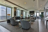 Located at 1792 Coventry Rd in Decatur, GA, this single-floor amenity space offers an elevated, contemporary environment designed for socializing and recreation. The layout flows seamlessly from a welcoming entryway into distinct functional zones, all unified by a cohesive modern aesthetic.

The entry foyer features tiled flooring, soft blue accent walls, and curated artwork, with glass doors opening to an outdoor terrace area filled with greenery. Recessed lighting sets a polished tone from the moment of arrival.

The lounge area is a standout feature, furnished with a dark sofa, swivel armchairs, and bold yellow leather ottomans arranged around a central coffee table. Floor-to-ceiling windows on multiple sides flood the space with natural light and frame impressive panoramic views of the surrounding cityscape. A wall-mounted television and fireplace add functionality and warmth to the seating zone.

Adjacent to the lounge, a bar area features a dark stone-topped counter with upholstered bar seating, overhead blue pendant lighting, and additional city views. The game room hosts a full-size pool table positioned beneath recessed ceiling lights, with direct access to the outdoor terrace through wide glass sliding doors. High-top tables and stools provide additional seating throughout. The overall ambiance is refined yet inviting, balancing comfort with upscale design.