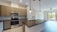 Located at 240 Grant St SE in Atlanta's vibrant Grant Park neighborhood, this single-floor residence offers a thoughtfully designed layout with modern finishes throughout. The kitchen serves as the centerpiece of the space, featuring sleek taupe shaker-style cabinetry extending to the ceiling, dark granite countertops, a full suite of stainless steel appliances including a French door refrigerator, electric range, built-in microwave, and dishwasher. A large kitchen island with an undermount sink and elegant pendant lighting above provides additional prep space and a natural gathering point. A subtle subway tile backsplash adds texture and dimension to the cooking area, while light wood-tone flooring ties the kitchen and living spaces together. The living area is bright and open, with a ceiling fan and sliding glass doors that lead to a private balcony overlooking lush greenery. The bedroom features plush carpeting and large windows that flood the room with natural light and outdoor views. The bathroom maintains the same design language as the rest of the home, with taupe cabinetry, dark granite countertops, a large vanity mirror, tile flooring, and a combined tub and shower. Clean white walls and recessed lighting throughout create a bright, contemporary ambiance.