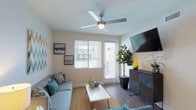This single-floor residence offers a thoughtfully designed layout with modern finishes and a cohesive teal and neutral color palette throughout. The living room serves as the central gathering space, featuring wood-look flooring, a ceiling fan, and a wall-mounted flat-screen TV positioned above a sleek striped media console. Natural light enters through blinds-fitted windows and a glass-paneled door that leads to an outdoor area. Comfortable seating, including a gray sofa and accent chair, is arranged around a hairpin-leg coffee table atop a natural jute rug.

The kitchen is well-equipped with rich dark wood cabinetry, granite countertops, a white glass tile backsplash, and a full suite of stainless steel appliances including a gas range, refrigerator, microwave, and dishwasher. A separate dining area is visible nearby.

The primary bedroom features carpeted flooring, a ceiling fan, a tufted upholstered headboard, and a window with outdoor views. A second bedroom offers a daybed with a trundle, bright windows, and carpet flooring.

Both bathrooms include wood-look tile flooring, tub-shower combinations, and clean white fixtures, with one bath featuring built-in open shelving for added storage.
