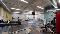 This fitness facility offers a spacious, open layout designed to accommodate a wide range of workout activities. The space features an industrial aesthetic, with exposed ductwork, structural support columns, and suspended fluorescent lighting running across the ceiling. The floors are covered in a patchwork of multi-toned carpet tiles in blue, grey, and tan, complemented by rubber mat sections in high-use workout zones.

The facility is divided into distinct activity areas. A cardio zone, set against yellow-painted walls, houses two treadmills and an elliptical trainer, each equipped with individual display monitors. A free weights area includes a dumbbell rack, a flat bench, and an open hex bar, while a separate section features a barbell bench press station and a standing punching bag for strength and boxing training. A cable machine station is positioned near a blue accent wall, which is decorated with motivational framed artwork. A wall-mounted flat-screen television provides entertainment throughout the main floor area.

Natural light enters through a row of windows along one wall, supplementing the overhead fluorescent fixtures. A water cooler station provides hydration access. White-painted concrete block walls and structural columns reinforce the utilitarian character of the space, creating a functional and practical environment suited for comprehensive fitness training.