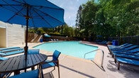 Located in Roswell, Georgia, this outdoor pool facility at Barrington Oaks Ridge offers a well-appointed recreational retreat designed for community enjoyment. The centerpiece is a generously sized swimming pool with clear, blue water, accessible via stainless steel handrails and marked with depth indicators for safety. The pool deck is constructed with a durable concrete surface that extends broadly around the perimeter, providing ample space for lounging and socializing. Furnished with multiple blue chaise lounges, outdoor tables, and chairs, the deck is thoughtfully arranged to accommodate both sunbathing and shaded relaxation under vibrant blue market umbrellas. A raised stone retaining wall with staircase access adds architectural interest and defines one boundary of the space, while a wooden deck section on the opposing side adds a warm textural contrast. An adjacent two-story clubhouse building with a covered porch overlooks the pool, providing a convenient facility anchor. The entire area is enveloped by tall, mature trees that offer natural shade, privacy, and a serene wooded ambiance. Safety features, including emergency call signage, are visibly integrated into the space. The overall atmosphere is bright, inviting, and well-maintained, making it an ideal outdoor amenity.