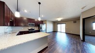 This single-floor space showcases an open-concept layout that seamlessly connects the kitchen and living area, creating a spacious and functional environment. The kitchen is appointed with rich dark-stained cabinetry that provides a striking contrast against the light granite countertops. A central island with matching granite extends the workspace and serves as a natural dividing element between the kitchen and living areas. Stainless steel appliances, including a refrigerator with ice maker, electric range, and microwave, are integrated throughout. Pendant lighting hangs above the island, complementing the recessed ceiling lights throughout the space. The living area is bathed in natural light from large sliding glass doors that open to a balcony, offering views of the surrounding exterior. Dark hardwood-style flooring runs continuously throughout both areas, adding warmth and visual cohesion to the space. Neutral-toned walls enhance the bright, airy atmosphere, while a textured ceiling adds a subtle design detail. The overall ambiance is modern and welcoming, well-suited for both everyday living and entertaining.