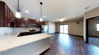 This single-floor space showcases an open-concept layout that seamlessly connects the kitchen and living area, creating a spacious and functional environment. The kitchen is appointed with rich dark-stained cabinetry that provides a striking contrast against the light granite countertops. A central island with matching granite extends the workspace and serves as a natural dividing element between the kitchen and living areas. Stainless steel appliances, including a refrigerator with ice maker, electric range, and microwave, are integrated throughout. Pendant lighting hangs above the island, complementing the recessed ceiling lights throughout the space. The living area is bathed in natural light from large sliding glass doors that open to a balcony, offering views of the surrounding exterior. Dark hardwood-style flooring runs continuously throughout both areas, adding warmth and visual cohesion to the space. Neutral-toned walls enhance the bright, airy atmosphere, while a textured ceiling adds a subtle design detail. The overall ambiance is modern and welcoming, well-suited for both everyday living and entertaining.