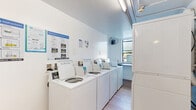 This community laundry facility on N 57th Ave in Hollywood, Florida offers a practical and well-organized space designed to serve residents efficiently. The single-floor layout is clean and bright, with white walls and light wood-style flooring that contribute to an open, tidy atmosphere. A row of top-load, coin-operated washing machines lines one wall, each equipped with digital payment interfaces and clearly posted operating instructions for user convenience. Stacked dryer units are positioned along the adjacent wall, maximizing the use of available space. A built-in countertop surface provides a dedicated area for folding laundry, while a wall-mounted fan ensures comfortable air circulation throughout the room. Natural light filters in through a window, adding to the overall brightness of the space. Safety is well addressed, with a mounted fire extinguisher, a ventilation panel, a sprinkler system, and clearly marked emergency exit signage and door. Overhead fluorescent lighting ensures the room remains well-lit at all times. The facility is accessible via a secured entry door and connects to an adjacent corridor, making it a convenient and functional amenity for the surrounding residential community.