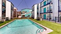Located at 4425 Gilbert Ave in Dallas, TX, this outdoor pool area offers a lively and well-appointed communal retreat. At the heart of the space is a generously sized rectangular swimming pool with clear turquoise water, lane markers, and depth indicators ranging from shallow to deep ends. The pool is surrounded by vibrant artificial turf that provides a clean, low-maintenance green landscape throughout the courtyard.

The space is enclosed by a multi-story residential building featuring a bold contemporary aesthetic — white exterior walls are accented by striking teal/turquoise doors and trim, with dark railings lining the balconies that overlook the pool area. A colorful mural on one of the building facades adds a dynamic artistic element to the environment.

String lights are strung overhead across the courtyard, creating an inviting ambiance for evening gatherings. A stone-accented bar or counter area with bench seating is positioned poolside, offering a functional social hub. Additional lounge chairs are arranged around the perimeter for relaxation and sunbathing.

The courtyard is secured by black iron fencing and includes pool safety signage. Mature palms and greenery punctuate the space, enhancing its resort-like feel. Open to the Dallas sky, the area benefits from abundant natural sunlight, making it an ideal outdoor amenity.