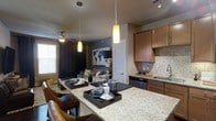 This one-bedroom, one-bathroom unit is designed with an open-concept floor plan that seamlessly connects the kitchen, dining, and living areas into a cohesive, functional space. The kitchen serves as a central focal point, featuring granite countertops with a large breakfast bar ideal for casual dining or entertaining. Warm-toned wooden cabinetry provides ample storage, while a decorative tile backsplash adds visual interest. Pendant lighting hangs above the bar, complementing the under-cabinet and track lighting to create a well-lit workspace. Stainless steel appliances and a stainless undermount sink complete the kitchen's polished look. The open living area beyond benefits from natural light streaming through a window fitted with blinds and draped with dark curtains that add contrast to the space. A ceiling fan ensures year-round comfort. The living area features a neutral color palette with an accent wall in deep charcoal, creating depth and a modern aesthetic. Overall, the unit balances contemporary style with practical design, offering a comfortable and visually cohesive living environment.
