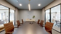 This single-floor space is thoughtfully designed to support both formal and informal meetings, combining polished aesthetics with functional layouts. The centerpiece is a sophisticated boardroom anchored by a long, dark conference table surrounded by cognac leather chairs with wooden frames. A striking geometric-patterned accent wall serves as a bold focal point, complemented by a sleek linear pendant light suspended from the gray ceiling. Recessed lighting and large windows dressed in floor-to-ceiling linen drapes allow natural light to filter into the space while maintaining privacy.

Adjacent to the boardroom, glass-paneled sliding doors with black frames connect to a casual lounge area, visible through the generous openings. This secondary space features comfortable gray sofas, built-in shelving with curated décor, and warm hardwood-style flooring, offering a relaxed atmosphere for informal gatherings. Patterned carpet flooring flows throughout the transitional corridors, adding texture underfoot. A glassed entrance with black-framed doors and sidelights provides a transparent, welcoming entry point. Together, the interconnected areas create a cohesive, modern environment ideal for professional use.