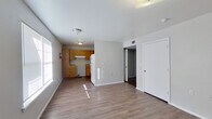 This single-story 2-bedroom, 1-bathroom residence offers a straightforward and functional layout across its single floor. The open living area is finished with light-toned wood-look flooring that extends throughout the home, lending a clean and cohesive look. Natural light filters in through the windows, brightening the neutral gray walls and white trim that define the interior palette.

The kitchen is positioned adjacent to the living space and features warm honey-toned wood cabinetry with ample upper and lower storage, a stainless steel sink, laminate countertops in an L-shaped configuration, and a ventilation hood above the range area. A white refrigerator completes the kitchen setup.

Both bedrooms are well-proportioned and share the same wood-look flooring and soft neutral wall tones. The primary bedroom offers a comfortable footprint and includes a double-door closet for storage. Each bedroom features at least one window fitted with blinds, allowing for natural light and privacy control.

The bathroom is equipped with a full-sized soaking tub and shower combination with a white surround, complemented by chrome hardware including a towel bar and toilet paper holder. A connecting hallway ties the bedrooms and bathroom together, creating an efficient and navigable floor plan throughout the home.