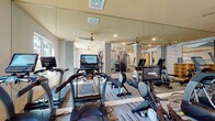 Located at 1027 Ashes Dr in Wilmington, NC, the Hudson Renaissance fitness center is a thoughtfully designed, single-floor facility built to support a wide range of workout routines. The main gym floor features a row of True-brand treadmills and cardio machines, each equipped with individual digital display screens for an interactive exercise experience. Floor-to-ceiling mirrors line the walls, visually expanding the space and allowing members to monitor their form during workouts. Recessed lighting and ceiling fans keep the environment bright and comfortable throughout. A dedicated strength training area showcases a True cable machine system positioned against a bold, geometric-patterned accent wall, adding a striking visual element to the functional space. Patterned, multi-toned carpet tiles cover the main floor, contributing to the gym's contemporary aesthetic. A separate studio room provides a more intimate setting for flexible training, featuring hardwood-style flooring, patterned wallcovering, wall-mounted mirrors, and a rack of exercise balls in vibrant colors. Glass-door entryways connect the spaces, maintaining an open and transparent flow. Natural light filters in through multiple windows, enhancing the overall welcoming and energetic atmosphere of the facility.