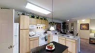 This single-floor space centers around a functional and well-appointed kitchen that combines practicality with a warm, inviting aesthetic. Light wood cabinetry lines the walls, providing ample storage that extends to the upper reaches, where decorative greenery adds a natural touch. The kitchen is equipped with white appliances, including a electric range with a ventilation hood, a built-in microwave, a dishwasher, and a refrigerator. A double sink is positioned along the perimeter cabinetry, offering a practical workspace. Countertops feature a speckled granite-style surface that complements the light cabinetry throughout. A striking dark-topped central island serves as a focal point, providing additional prep and casual dining space, and is accented by a pendant light that drops from the ceiling. Overhead, a large skylight-style fluorescent fixture brightens the workspace, while track lighting adds additional illumination. The kitchen opens naturally into a dining area, visible in the background, where a framed artwork and lush indoor plants create a warm transition between spaces. Large windows in the adjoining area allow natural light to filter through, enhancing the overall brightness of the space.