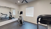 This dedicated fitness space is thoughtfully designed to support a comprehensive workout routine within a clean, functional environment. The room features durable rubber flooring throughout, providing a safe and resilient surface ideal for exercise equipment and high-impact activity. Crisp white walls and recessed ceiling lighting create a bright, energizing atmosphere, while a ceiling fan ensures consistent airflow and comfort during workouts. A large wall-mounted mirror spans one side of the room, enhancing the sense of space and allowing users to monitor their form during exercise. Natural light filters in through windows fitted with horizontal blinds, adding to the room's inviting feel. The space is equipped with a diverse selection of fitness machines, including a treadmill, elliptical trainer, recumbent stationary bike, and a multi-station cable strength machine — offering a well-rounded combination of cardio and resistance training options. A dedicated panel-style door provides easy access to and from the room. The overall layout is efficient and purposeful, making this fitness room a practical and complete facility for daily training needs.