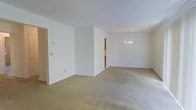 This single-floor space presents an open and airy interior characterized by clean white walls and wall-to-wall neutral beige carpeting throughout. The layout flows seamlessly between connected areas, with a partial wall dividing the space into distinct functional zones while maintaining an open feel. A notable architectural feature is the elegant chandelier suspended from the ceiling in the far room, adding a refined focal point to what appears to be a dining or secondary living area. The ceilings are standard height and painted white, contributing to the overall bright and neutral atmosphere. Doorways visible along the walls provide access to additional rooms or hallways, suggesting a well-connected floor plan. A thermostat is mounted on one wall, and standard electrical outlets are positioned throughout. The absence of furniture allows the full extent of the carpeted floor space to be appreciated, highlighting the generous square footage and versatility of the layout. The uniformly white walls and light-toned carpet create a blank canvas well-suited for a variety of interior design styles.