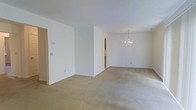 This single-floor space presents an open and airy interior characterized by clean white walls and wall-to-wall neutral beige carpeting throughout. The layout flows seamlessly between connected areas, with a partial wall dividing the space into distinct functional zones while maintaining an open feel. A notable architectural feature is the elegant chandelier suspended from the ceiling in the far room, adding a refined focal point to what appears to be a dining or secondary living area. The ceilings are standard height and painted white, contributing to the overall bright and neutral atmosphere. Doorways visible along the walls provide access to additional rooms or hallways, suggesting a well-connected floor plan. A thermostat is mounted on one wall, and standard electrical outlets are positioned throughout. The absence of furniture allows the full extent of the carpeted floor space to be appreciated, highlighting the generous square footage and versatility of the layout. The uniformly white walls and light-toned carpet create a blank canvas well-suited for a variety of interior design styles.