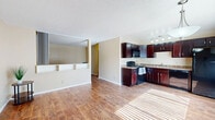 This single-floor unit offers a well-organized and efficient layout that makes smart use of its compact footprint. The combined kitchen and living area forms the heart of the space, creating an open, connected feel throughout. The kitchen is well-appointed with rich dark mahogany cabinetry extending to the upper walls, granite-style countertops, and a full suite of black appliances including a refrigerator, dishwasher, electric range, and built-in microwave. A track lighting fixture above the kitchen provides focused illumination, complementing the natural light that filters through the windows. The living area flows naturally from the kitchen and features a distinctive built-in half-wall partition with open shelving below, adding architectural character and a subtle sense of division between spaces. Warm hardwood-style flooring runs consistently throughout the unit, contributing to a cohesive and inviting ambiance. Neutral beige walls and a textured ceiling complete the interior aesthetic. A dedicated closet provides practical storage. The overall layout is straightforward and functional, making the most of every square foot while maintaining a clean, livable atmosphere.