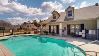 Located in Hammond, Louisiana, this residential community pool area offers a welcoming outdoor retreat designed for relaxation and recreation. The centerpiece is a generously sized swimming pool with clear, blue-green water, framed by a wide concrete sundeck lined with lounge chairs — ideal for soaking up the Louisiana sunshine. The pool area is fully enclosed by a black wrought iron fence, providing both security and a polished aesthetic, with access restricted to residents and their guests via a pool pass system.

Adjacent to the pool is a charming single-story clubhouse building with a traditional Southern architectural style, featuring dormered rooflines, a neutral stucco and siding exterior, and a covered porch supported by classic white columns. The porch is adorned with potted ferns and greenery, adding a warm, inviting touch. French-style glass doors with louvered shutters serve as the main entrance, welcoming visitors with a "Welcome Home" mat at the threshold.

A covered walkway connects the surrounding residential units to the pool area, featuring a wood-paneled ceiling and concrete flooring. Mature trees and well-maintained landscaping frame the property, while an American flag flies prominently near the pool deck, reinforcing the community's welcoming character. The overall ambiance is clean, cheerful, and community-oriented.