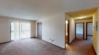 This single-floor residential unit offers a practical and comfortable layout with neutral-toned interiors throughout. The spacious living area is finished with wall-to-wall carpeting and features a textured ceiling, while large vertical-blind-covered sliding glass doors open to an outdoor balcony, filling the room with natural light and providing views of surrounding trees. A thermostat on the wall adds convenience for climate control.

The kitchen is equipped with warm oak cabinetry offering ample upper and lower storage, a double-basin sink, and a light-toned countertop. White appliances include a refrigerator, dishwasher, electric range, and oven. The flooring in the kitchen features durable tile in a neutral pattern.

The bedrooms are carpeted and bathed in natural light through windows that frame peaceful wooded views. Each room provides a calm, functional space with clean walls and dark wood baseboards that complement the overall aesthetic.

The bathroom includes a full tub-and-shower combination with white surround, a toilet, and a vanity with an oak cabinet base and a mirror above. A secondary vanity area with additional under-sink storage is accessible from the hallway, accompanied by a sliding mirrored closet door. Tile flooring runs throughout the wet areas, maintaining a clean and practical finish across the unit.