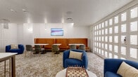This thoughtfully designed single-floor amenity space blends social, functional, and recreational areas into one cohesive environment. The interior is anchored by a warm, neutral palette of whites and natural wood tones, accented by gold hardware and patterned flooring that flows throughout the space.

A standout feature is the plush, cognac-colored leather banquette running along a white paneled wall, paired with white-topped tables and velvet dining chairs with gold legs — an inviting area for casual meals or social gatherings. A wall-mounted flat-screen TV and elegant pendant lighting add ambiance to this seating zone.

Adjacent to the lounge seating is a dedicated workspace area, centered around a large white conference-height table surrounded by upholstered chairs, ideal for collaborative work or meetings. A large glass whiteboard panel nearby enhances the space's productivity-focused functionality.

The wet bar area features warm wood open shelving with diagonal wine storage, lower cabinetry, a sink, and counter seating — making it well-suited for entertaining or informal gatherings. A tray ceiling with recessed lighting adds a refined architectural detail overhead.

A relaxed lounge nook hosts two plush blue accent chairs around a marble-top coffee table set with a decorative chess board. A full wall of white package lockers provides a practical, organized storage solution, completing this versatile and well-appointed amenity space.