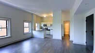 This single-floor space showcases a clean, modern aesthetic with an open-concept layout that maximizes the sense of space and ease of movement throughout. Dark hardwood flooring extends across the main living area, providing a striking contrast against the crisp white walls and light-toned ceilings.

The kitchen is thoughtfully designed with white cabinetry, a breakfast bar peninsula, and stainless steel appliances including a range and over-the-range microwave. Natural light filters in through multiple windows, brightening the open living and kitchen areas and creating a welcoming, airy atmosphere.

Structural columns are integrated into the layout, adding an architectural element while subtly defining the transition between the living space and kitchen. The open sightlines from the living area through to the kitchen make the space feel expansive and connected.

A hallway leads to additional areas of the unit, with a glimpse of a bathroom visible in the background, suggesting a well-organized and functional floor plan. The overall ambiance is clean, contemporary, and move-in ready, with neutral tones and quality finishes that provide a versatile backdrop for a variety of interior styles.