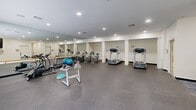Located at 1800 Palmer Rd in Fort Washington, Maryland, this fitness center offers a single-floor exercise room designed to support a well-rounded workout routine. The space features durable rubber flooring throughout, providing both safety and comfort during exercise. Recessed lighting illuminates the room evenly, complemented by natural light streaming in through a row of large windows that offer views of surrounding greenery, creating an open and inviting atmosphere. A full-length mirrored wall with a ballet barre runs along one side of the room, visually expanding the space and serving as a functional feature for stretching and form-checking. The fitness room is equipped with a variety of cardio machines, including treadmills, stationary bikes, and an elliptical trainer. Free weights, kettlebells, exercise balls, and a dumbbell rack provide ample options for strength and functional training. A wall-mounted flat-screen television adds entertainment for those engaged in longer cardio sessions. A large standing fan supports air circulation within the space. The room also includes accessible entry points, with signage indicating ADA-compliant facilities. Clean white walls and a neutral color palette contribute to the bright, functional ambiance of this well-appointed fitness facility.