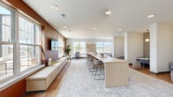 Located at 8681 Edison Plaza Dr in Fishers, Indiana, this thoughtfully designed community lounge space offers a versatile, single-level open-plan layout. The interior is defined by a warm blend of natural wood tones, crisp white walls, and rich walnut wood-paneled accent walls that anchor the space with a sophisticated, contemporary character.

At the heart of the room sits a long, light wood communal bar-height table with modern bar stools, centered on a textured area rug and ideal for collaborative gatherings or casual dining. Recessed ceiling lighting and large multi-pane windows along multiple walls flood the interior with natural light while offering outdoor views of the surrounding plaza and courtyard.

The living area transitions seamlessly into distinct zones: a lounge corner features plush upholstered accent chairs, a fiddle-leaf fig plant, and wall-mounted decorative art; cozy semi-private nook seating areas are set against geometric-patterned wallpaper with elegant globe pendant lights, offering intimate booth-style tables. A low-profile credenza runs along the wood-paneled wall, complemented by a wall-mounted display screen. Warm hardwood flooring flows throughout, grounding the bright, airy atmosphere. The overall design balances communal functionality with refined, welcoming aesthetics.