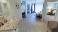 This single-floor residence showcases a thoughtfully designed open-concept layout that effortlessly connects the kitchen, dining, and living areas, creating a spacious and cohesive environment. The kitchen features elegant granite countertops with warm tones and speckled detailing, providing a refined anchor to the entertaining space.

Smooth, light-toned wood-look flooring extends throughout the entire living area, lending a clean, contemporary aesthetic and a sense of continuity. The living room is generously sized and bathed in natural light, thanks to large windows dressed with vertical blinds and dark curtain panels that offer both style and privacy. A ceiling fan with a classic fixture provides both comfort and subtle visual interest overhead.

Built-in shelving niches along the walls offer practical display space, adding character to the otherwise clean-lined interior. Neutral gray-toned walls create a versatile backdrop, complementing a range of furnishing styles. The open flow of the space makes it ideal for both everyday living and entertaining, while the abundance of natural light contributes to an airy, welcoming ambiance throughout. Overall, the residence combines modern finishes with a functional, open layout for comfortable single-floor living.
